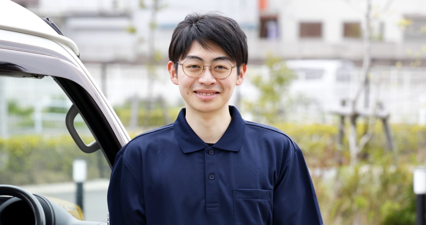 担当する軽バンの運転席ドアの横で、眼鏡をかけ穏やかに微笑むS.S.さん