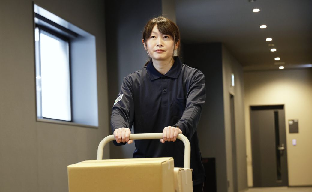 屋内の廊下にて、台車に載せた段ボール箱を運びながら前方を見つめるY.K.さん