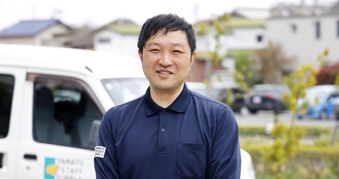 住宅街の柔らかな光の中で、配送車両（軽バン）を背景に柔和な表情で微笑むA.H.さん