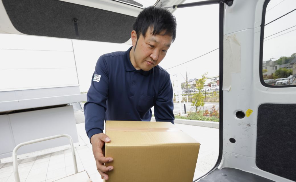 配送車両（軽バン）の荷台から、両手で段ボール箱をしっかり抱えて運び出そうとするA.H.さん