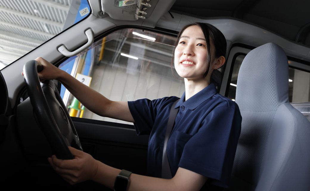 軽バン車両の運転席でハンドルを握り、明るい表情で前方を見つめるM.T.さん