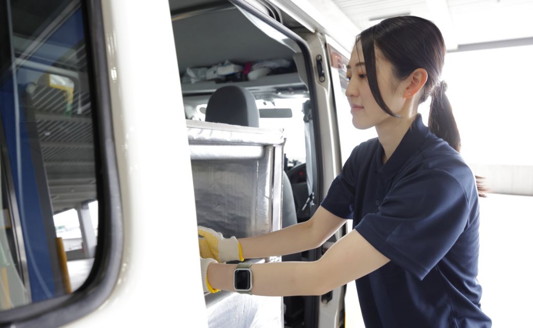 配送車両（軽バン）のスライドドアを開け、荷台に積まれた大きな保冷バッグへ手を伸ばすM.T.さん